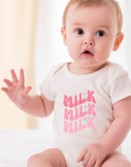 Baby wearing a white onesie with pink text Milk Milk Milk, sitting on a white surface.