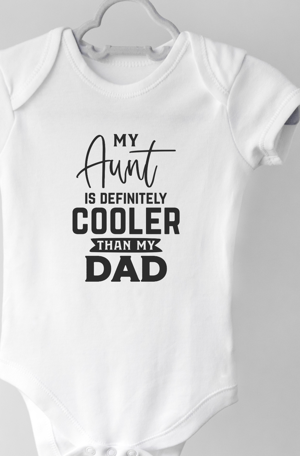 White baby onesie with text "My aunt is definitely cooler than my dad" on a hanger against a white backgrounds 