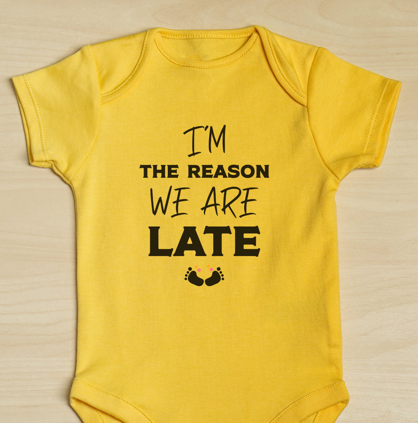 Yellow baby onesie with text I'm the reason we are late on a wooden surface
