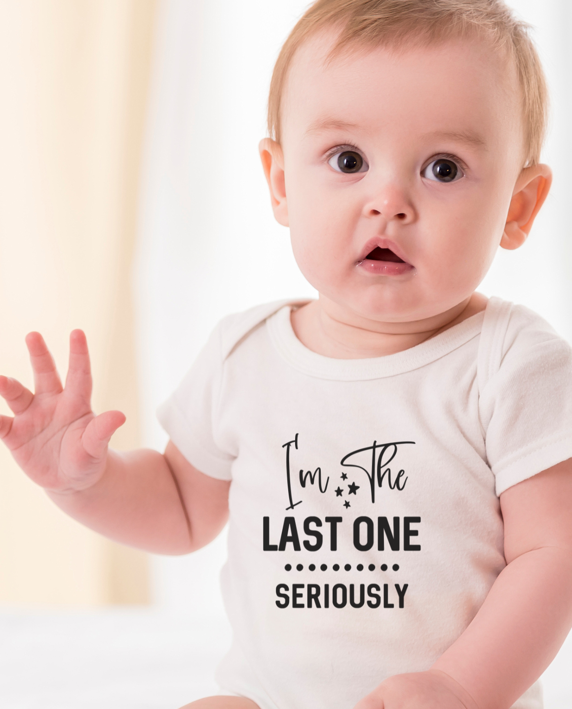 Baby wearing a white onesie with text " I'm the last one" sitting on a white surface.