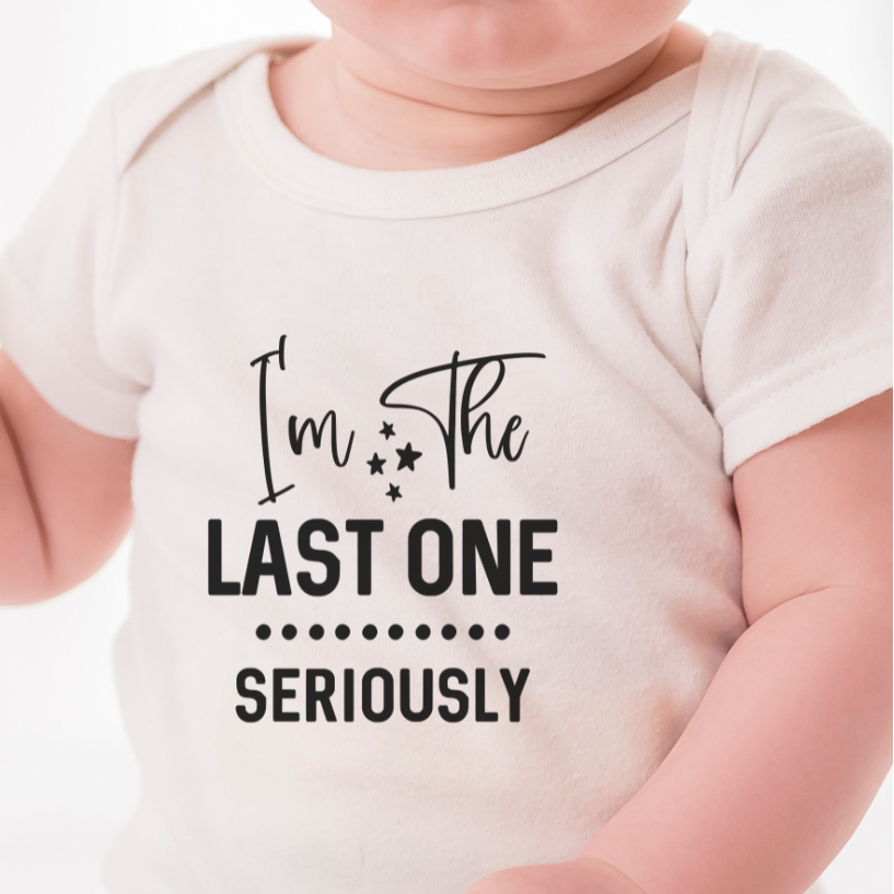 Baby wearing a white onesie with text " I'm the last one" sitting on a white surface.