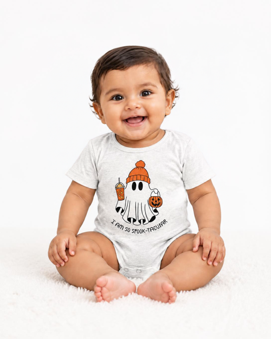 A smiling baby sitting on a white fluffy rug wearing a white short-sleeve bodysuit with a cartoon ghost graphic holding an orange pumpkin and drink, with the text 'I CAN DO TOO-TROUBLE' below.
