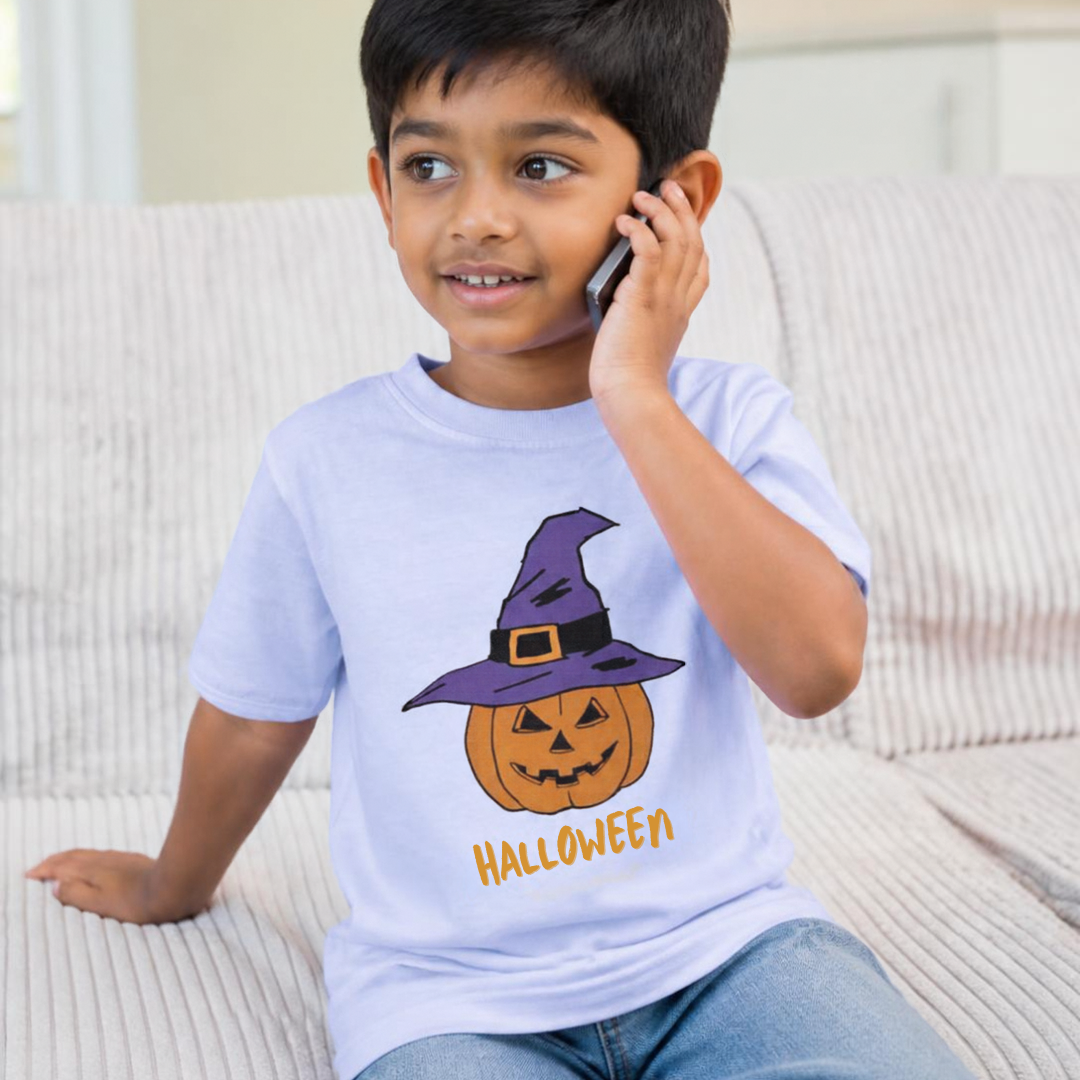 A young boy sitting on a light-colored couch, wearing a light blue t-shirt with a cartoon pumpkin wearing a purple witch hat and the word 'HALLOWEEN' printed on the front, while talking on a mobile phone.