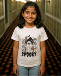 A young girl with dark hair stands smiling in a hotel hallway wearing a white graphic t-shirt that says 'SOCKTY SPOOKY' with a ghost and bat design, paired with light blue jeans.