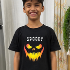 A young boy smiling while wearing a black t-shirt with a 'SPOOKY' text graphic and a yellow, orange, and red jack-o'-lantern face, standing in a home environment with curtains and a refrigerator in the background.