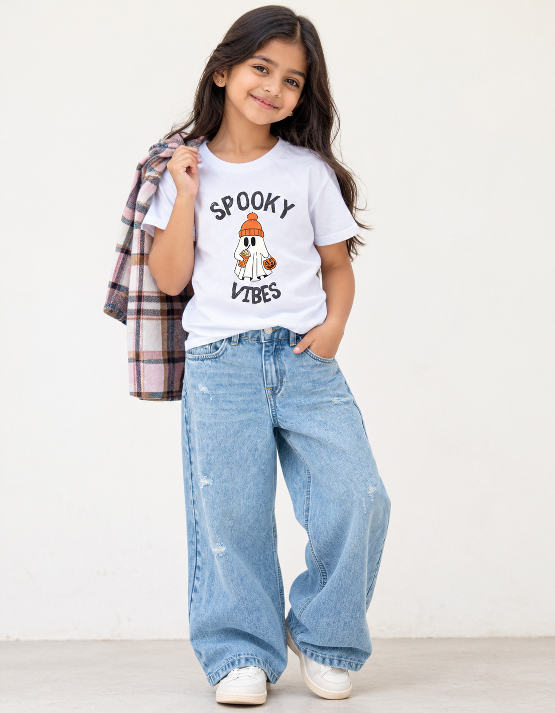 A young girl modeling a white graphic t-shirt with a cartoon ghost and the text 'SPOOKY VIBES', paired with wide-leg light blue jeans and holding a plaid shirt over her shoulder, set against a plain off-white wall.