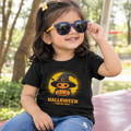 A young child wearing a black graphic t-shirt with a cartoon jack-o'-lantern in a witch hat and the text 'HALLOWEEN' on the front, paired with blue jeans and purple sunglasses, sitting outdoors.