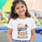A young girl with dark hair and a pink hair clip is smiling, wearing a white graphic t-shirt with a Halloween-themed design that reads 'When Witches Go Riding, Black Cats Love the Moon, It's Not Halloween' in orange and black lettering. The background is a blurred outdoor play area with colorful structures.