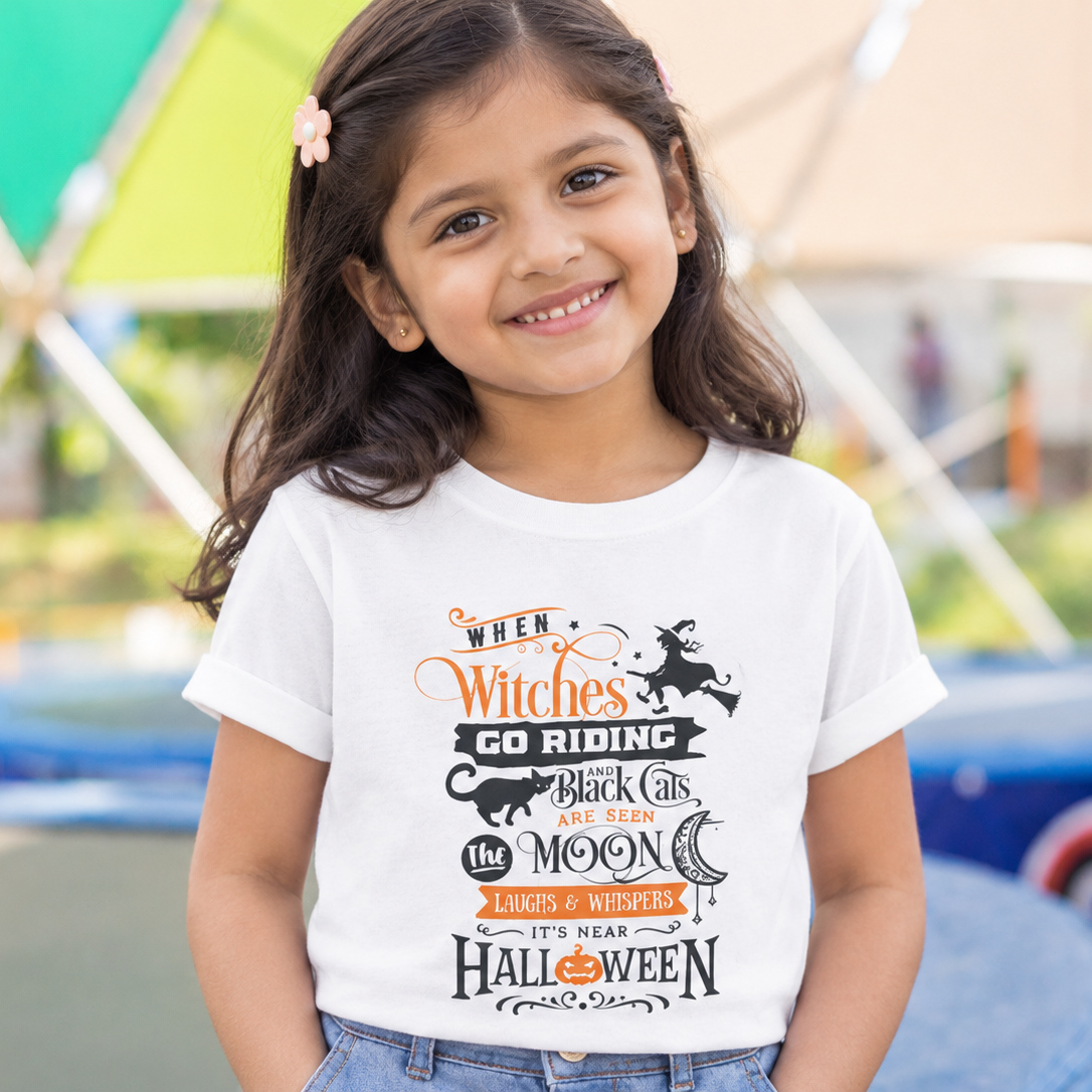 A young girl with dark hair and a pink hair clip is smiling, wearing a white graphic t-shirt with a Halloween-themed design that reads 'When Witches Go Riding, Black Cats Love the Moon, It's Not Halloween' in orange and black lettering. The background is a blurred outdoor play area with colorful structures.
