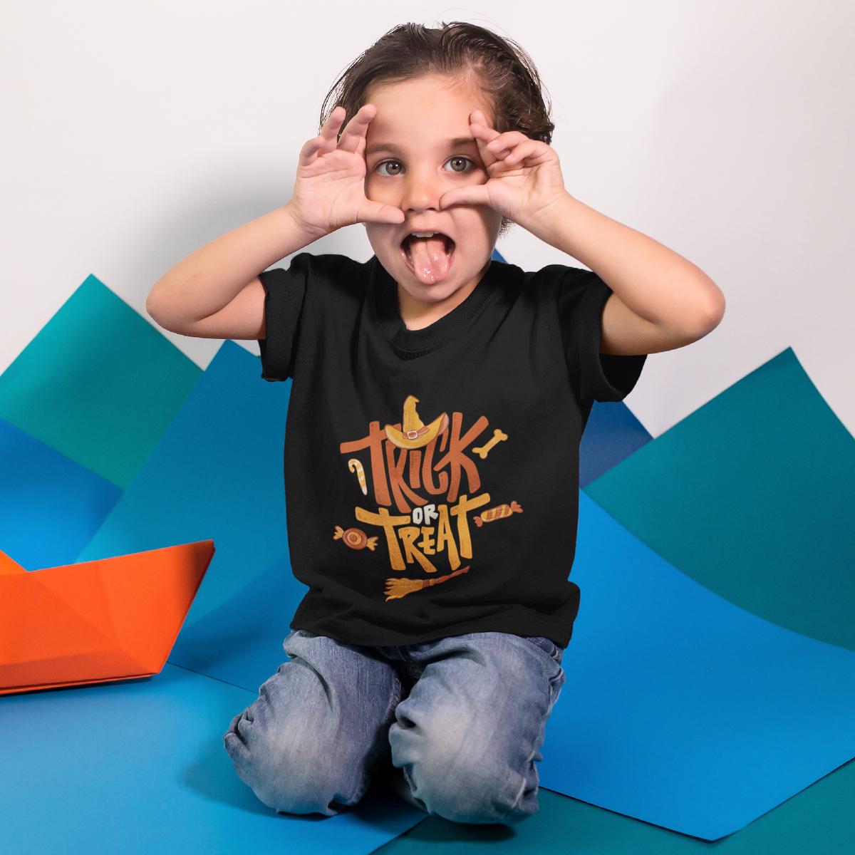Child wearing a black t-shirt with 'Trick or Treat' text, sitting on colorful paper boats.