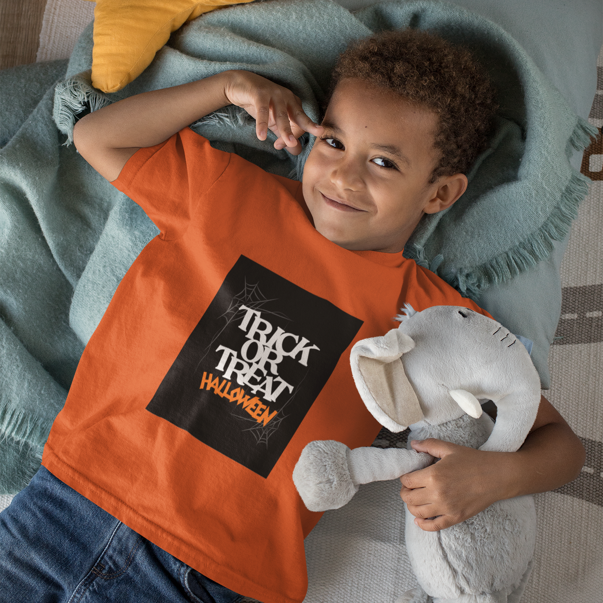 Child wearing an orange 'Trick or Treat Halloween' shirt, holding a stuffed elephant toy, on a patterned rug.