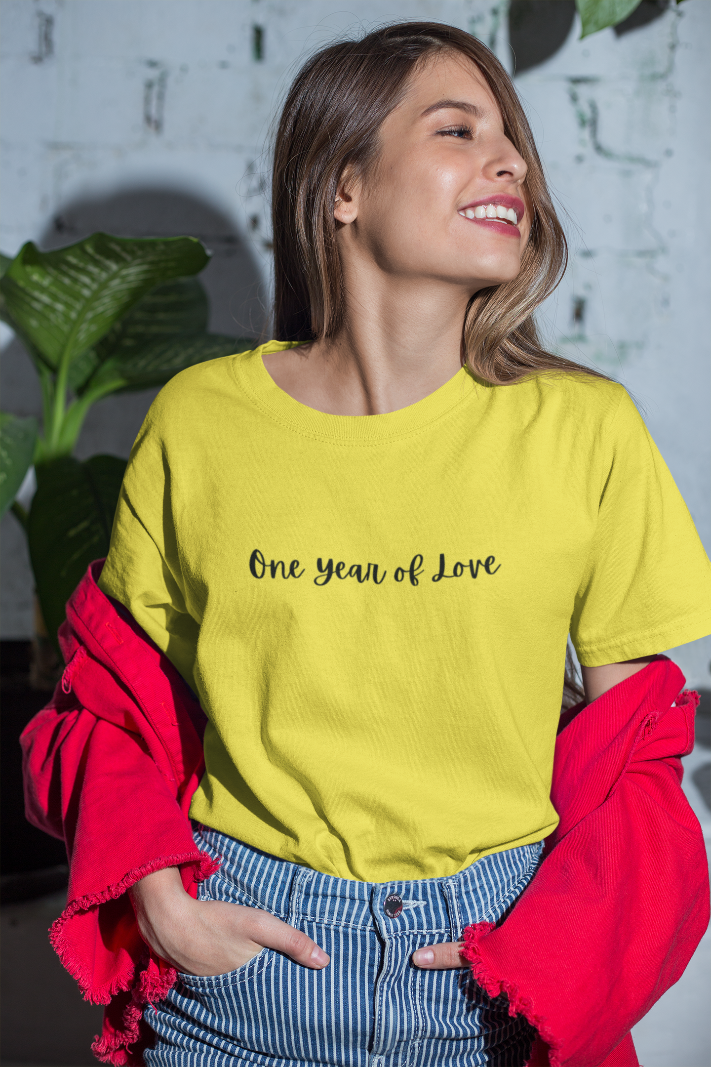 Woman wearing a yellow t-shirt with 'One Year of Love' text, standing indoors with plants in the background.